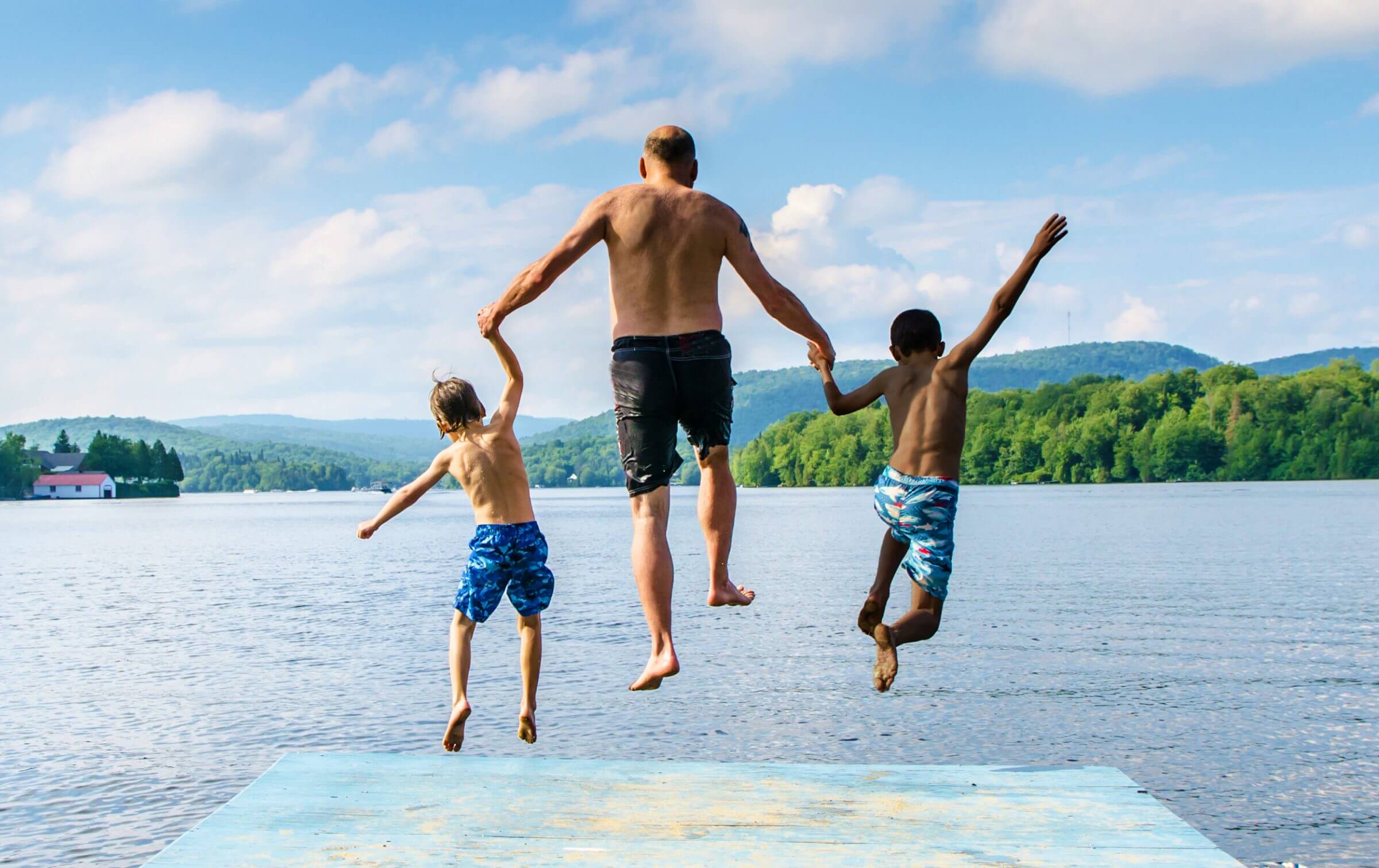 Photo from behind of a grandfather jumping off a dock into a lack with grandchildren at either side, arms raised in mid air.