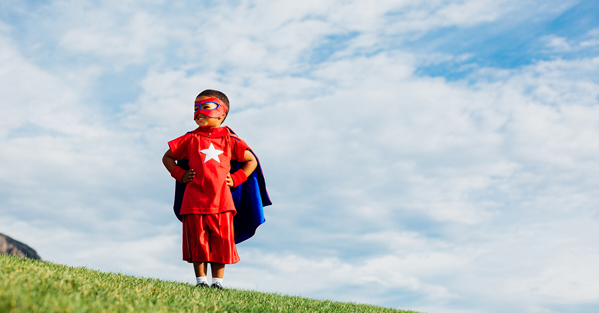 Young boy standing on a grassy hill with sky in the background. He is smiling, wearing a super hero custume with hands on hips.